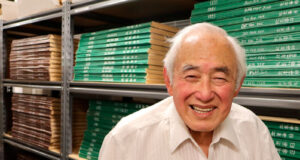 Smiling elderly man in a white shirt stands in a storage room with shelves of green and brown boxes.