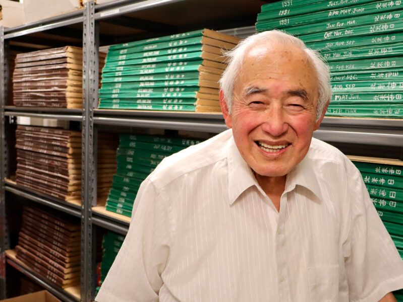 Smiling elderly man in a white shirt stands in a storage room with shelves of green and brown boxes.