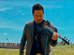 Man in a suit playing a violin outdoors in a grassy field under a bright blue sky