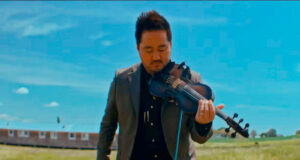 Man in a suit playing a violin outdoors in a grassy field under a bright blue sky