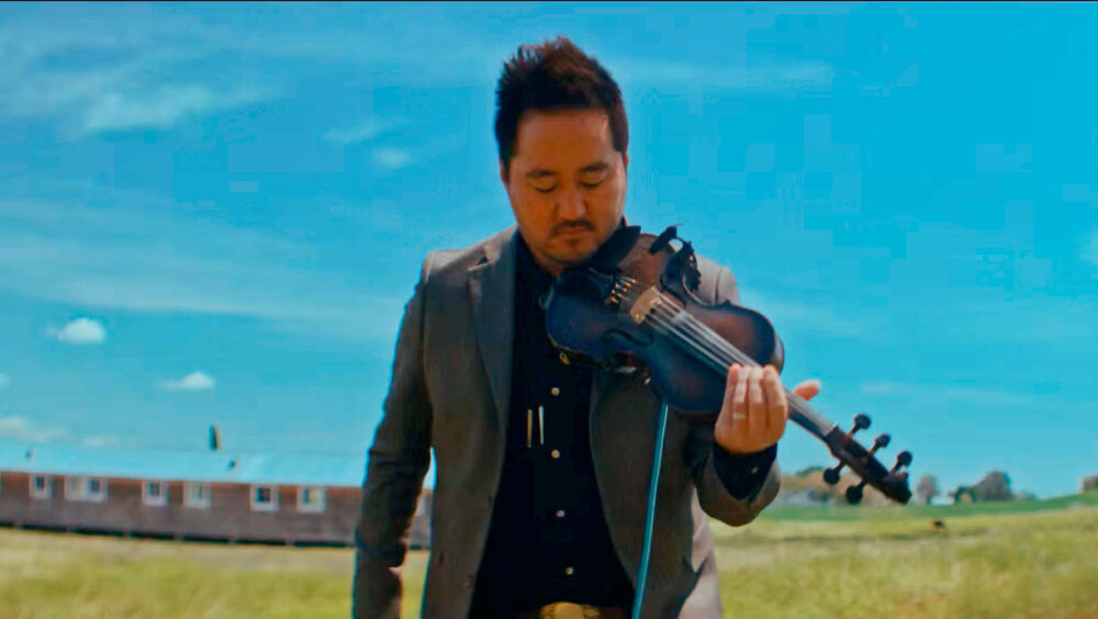 Man in a suit playing a violin outdoors in a grassy field under a bright blue sky