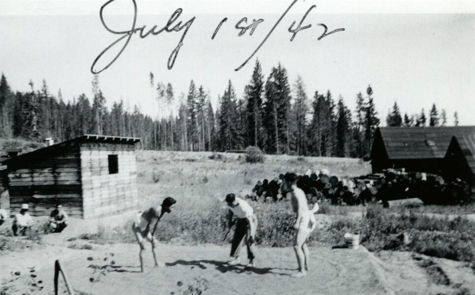 Black-and-white photo of children playing a game on a dirt field at a rural camp, with a wooden shack and cabins in the background and trees beyond.