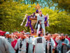 Festival procession: people in traditional robes carry a decorated portable shrine (mikoshi) through a sunny park, with a performer standing on top guiding the parade.