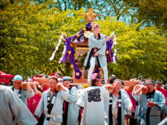 Festival procession: people in traditional robes carry a decorated portable shrine (mikoshi) through a sunny park, with a performer standing on top guiding the parade.