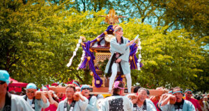 Festival procession: people in traditional robes carry a decorated portable shrine (mikoshi) through a sunny park, with a performer standing on top guiding the parade.
