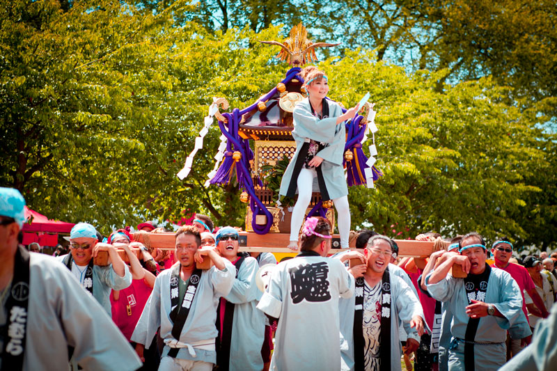 Festival procession: people in traditional robes carry a decorated portable shrine (mikoshi) through a sunny park, with a performer standing on top guiding the parade.