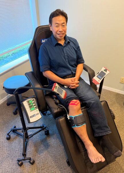 Man in a blue shirt sits in a specialized therapy chair, with a leg compression/cooling device strapped to his right leg and connected control panels nearby.
