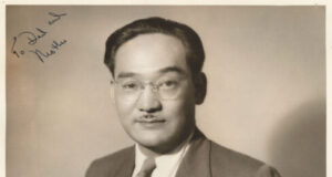 Portrait of a man in a suit and glasses, standing with hands in pockets, posing for a formal studio photo; handwritten notes appear in the corners.