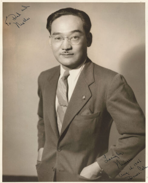 Portrait of a man in a suit and glasses, standing with hands in pockets, posing for a formal studio photo; handwritten notes appear in the corners.