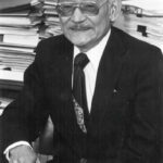 Older man in a suit with a tie and glasses, seated at a desk with stacks of papers in the background, holding a stamp or small object.