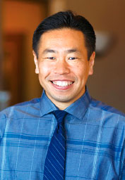 Smiling man in a blue plaid shirt and navy tie photographed indoors.