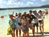 Okinawan Culture, Sunshine, Baseball and More in February 2027! Group of eight people posing playfully in shallow tropical water at a beach, smiling and waving at the camera.