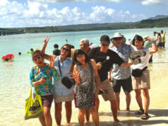 Group of eight people posing playfully in shallow tropical water at a beach, smiling and waving at the camera.