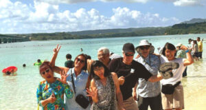 Group of eight people posing playfully in shallow tropical water at a beach, smiling and waving at the camera.