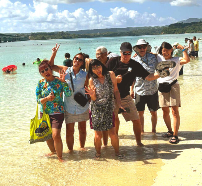 Group of eight people posing playfully in shallow tropical water at a beach, smiling and waving at the camera.