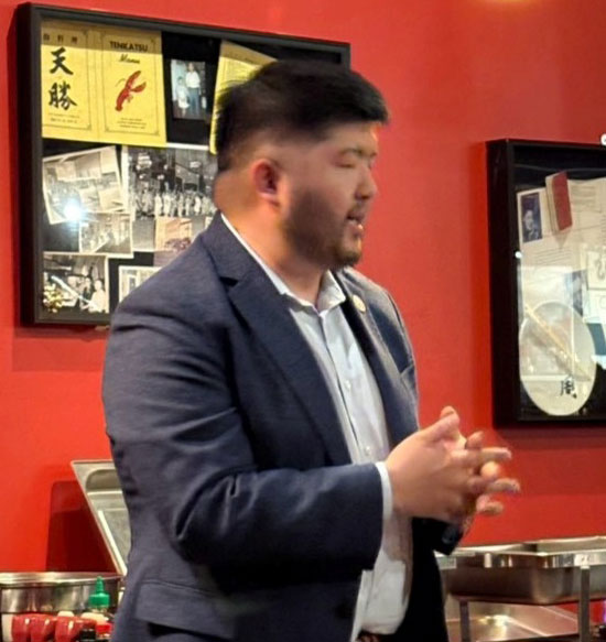 Side profile of a man in a dark blue blazer and white shirt speaking with hands clasped, in a red-walled room with framed photos on display.