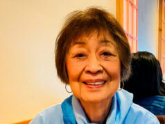 Ahead Of Her Time – Dolores Sibonga Smiling older woman in a light blue shirt with a blue scarf tied at the neck, sitting at a table in a warm-colored room.
