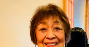 Smiling older woman in a light blue shirt with a blue scarf tied at the neck, sitting at a table in a warm-colored room.