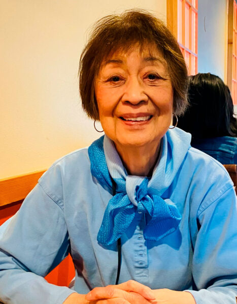 Smiling older woman in a light blue shirt with a blue scarf tied at the neck, sitting at a table in a warm-colored room.