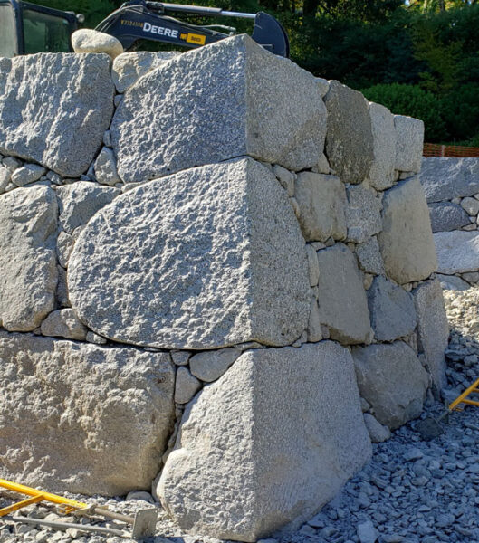 Rocky stone retaining wall under construction with large granite blocks and a blue excavator on top.