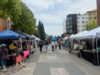 Outdoor street market with white vendor tents lining both sides; people browse stalls and chat on a sunny day.