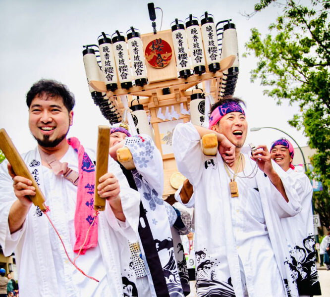 People in white festival robes smiling as they carry a wooden float adorned with lanterns during a Japanese festival.