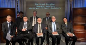 Nisei Paradox: Japanese American Draft Resisters during World War II Group of eight men in suits posing for a panel photo on stage, with a screen reading 'The Nisei Paradox: Japanese American World War II Draft Resisters' behind them, and flags in the background.