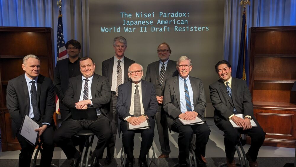 Group of eight men in suits posing for a panel photo on stage, with a screen reading 'The Nisei Paradox: Japanese American World War II Draft Resisters' behind them, and flags in the background.