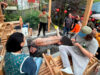 Group of people watching a pig roasting on a spit over an open charcoal pit at an outdoor gathering.