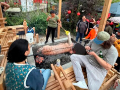 51st Annual Pig Roast Thank You! Group of people watching a pig roasting on a spit over an open charcoal pit at an outdoor gathering.