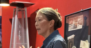 Woman speaking and gesturing with hands in a red-walled room, giving a presentation or talk.