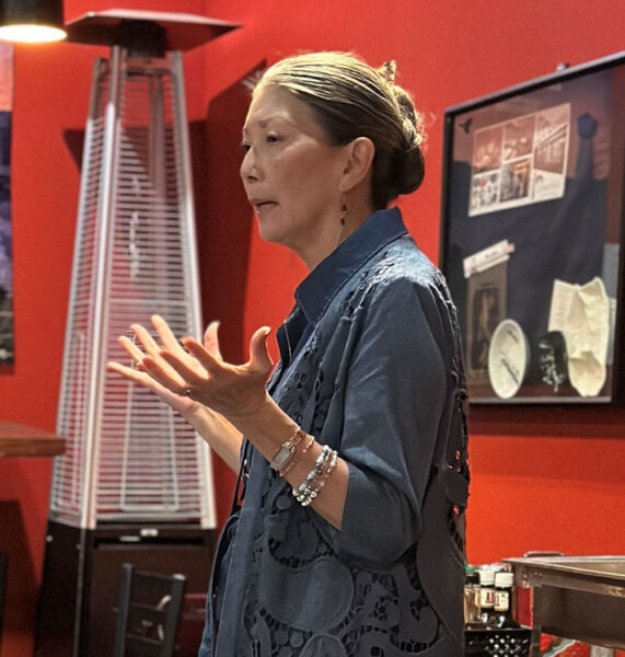 Woman speaking and gesturing with hands in a red-walled room, giving a presentation or talk.